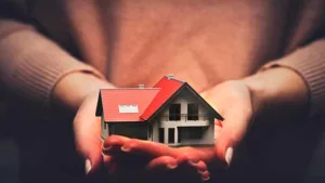 Hands cupping a small model house with a red roof, symbolizing home protection or real estate care.